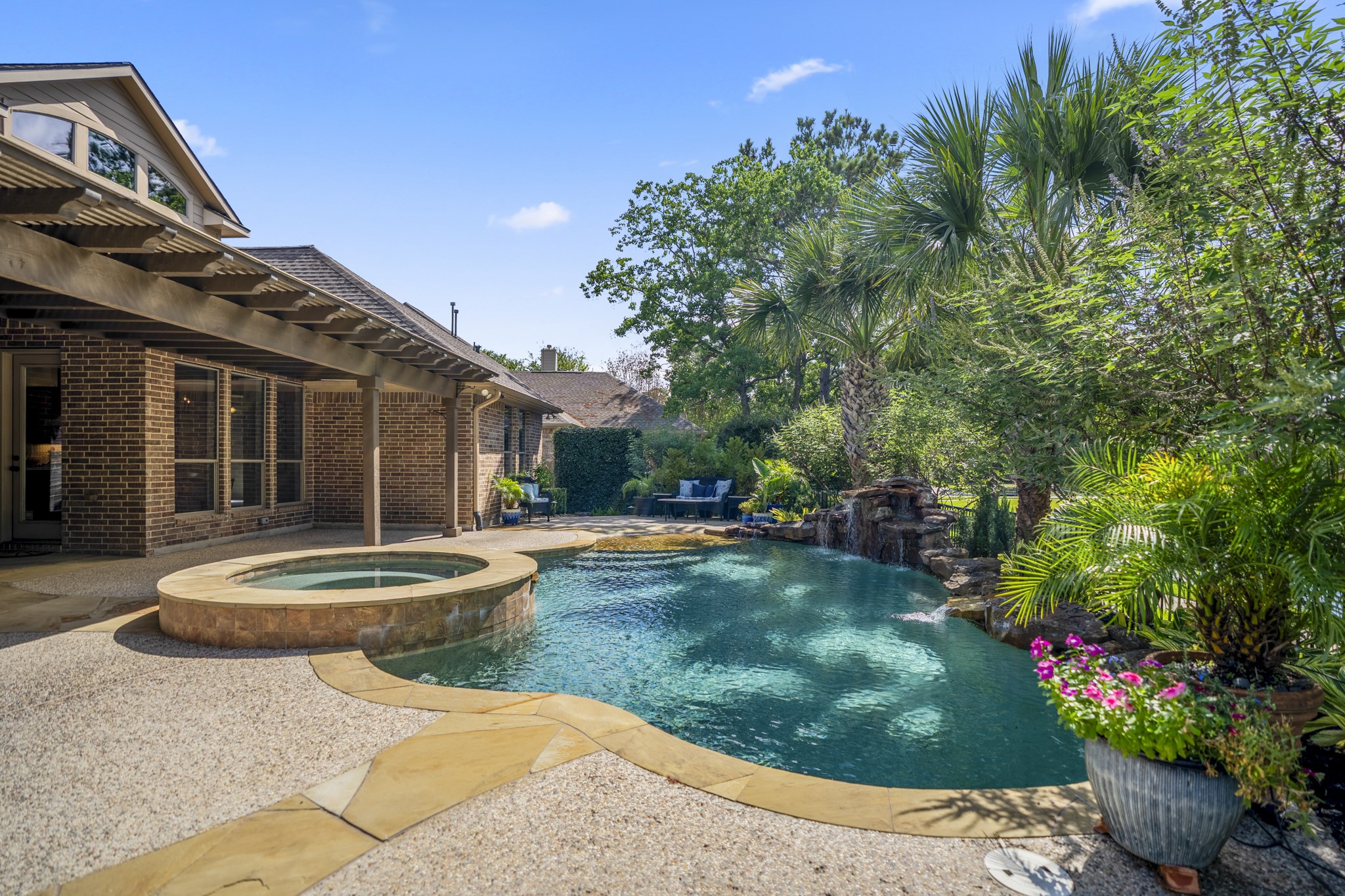 2407 Hendricks Lakes Drive Spring, TX 77388 - Photo 38 of 50 Poolside, the backyard feels wrapped in greenery, with the raised spa, curved pool, and surrounding landscaping creating a private-feeling place to relax outdoors. The covered pergola stretches along the back of the home on the left, while palm trees and layered shrubs soften the view and set up the kind of evening backdrop the sellers loved most.