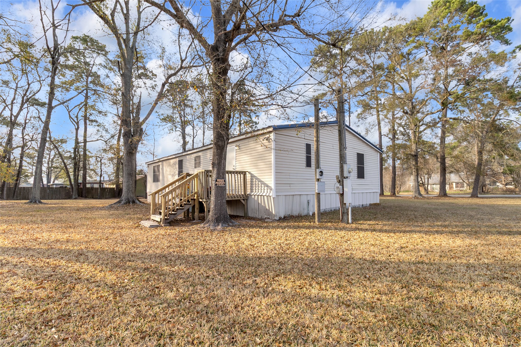 22094 Spear Road Porter, TX 77365 - Photo 4 of 45 This photo shows a mobile home situated on a spacious, grassy lot with mature trees. The exterior is simple, featuring a small wooden entry backdoor deck. Ideal for those seeking tranquility and outdoor space.