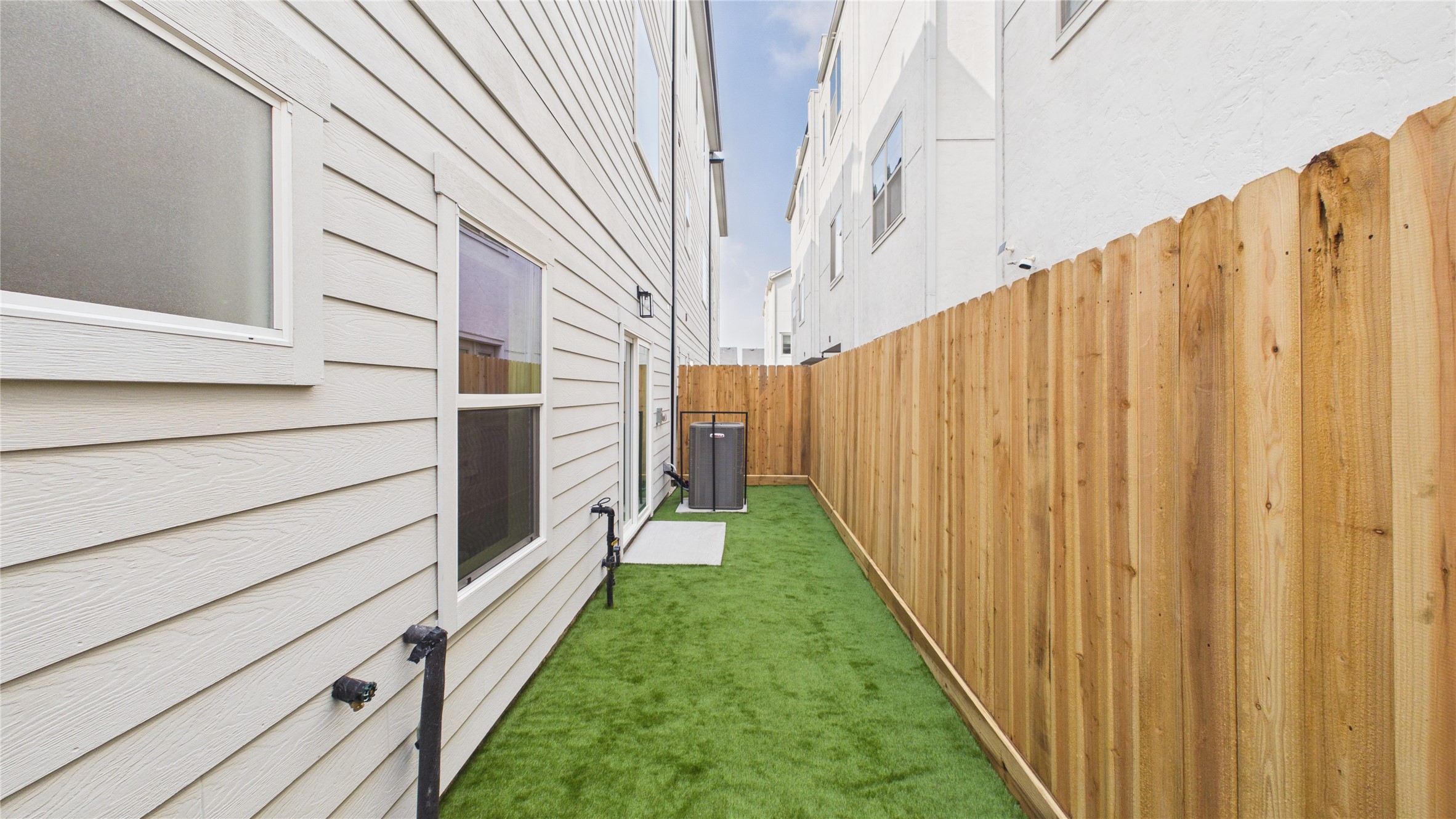 602 Skyland Terrace Way Houston, TX 77009 - Photo 37 of 40 This photo shows a narrow side yard with artificial grass between two buildings. It features a wooden fence on one side and the exterior wall of a house with windows on the other. An air conditioning unit is visible at the end of the yard.