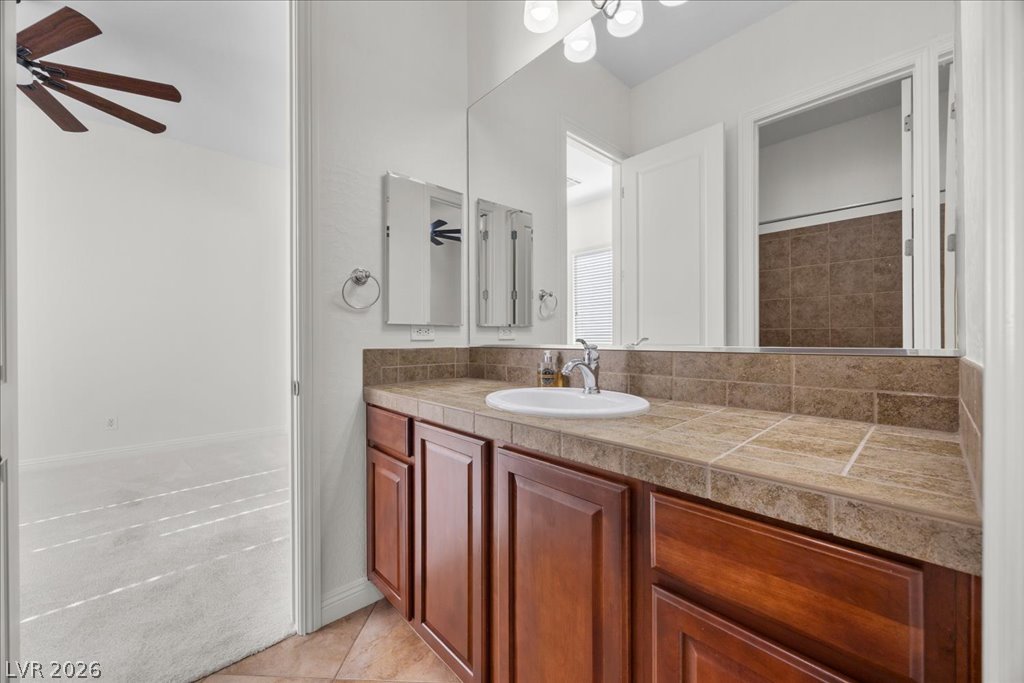 2125 De Rouge Avenue Henderson, NV 89044 - Photo 56 of 96 A well-appointed bathroom vanity features a tile countertop and rich wood cabinetry, with a mirrored view into the adjacent shower.