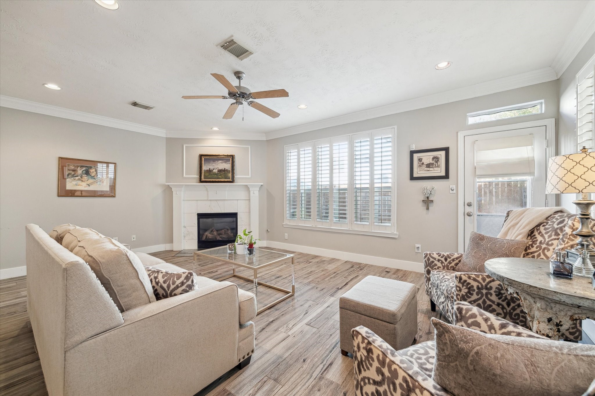 5242 Eigel Street Houston, TX 77007 - Photo 5 of 29 This inviting living room features a cozy fireplace, neutral walls, and wood-style flooring. It’s bright and airy with large windows and white shutters, complemented by a ceiling fan and recessed lighting for a modern touch.