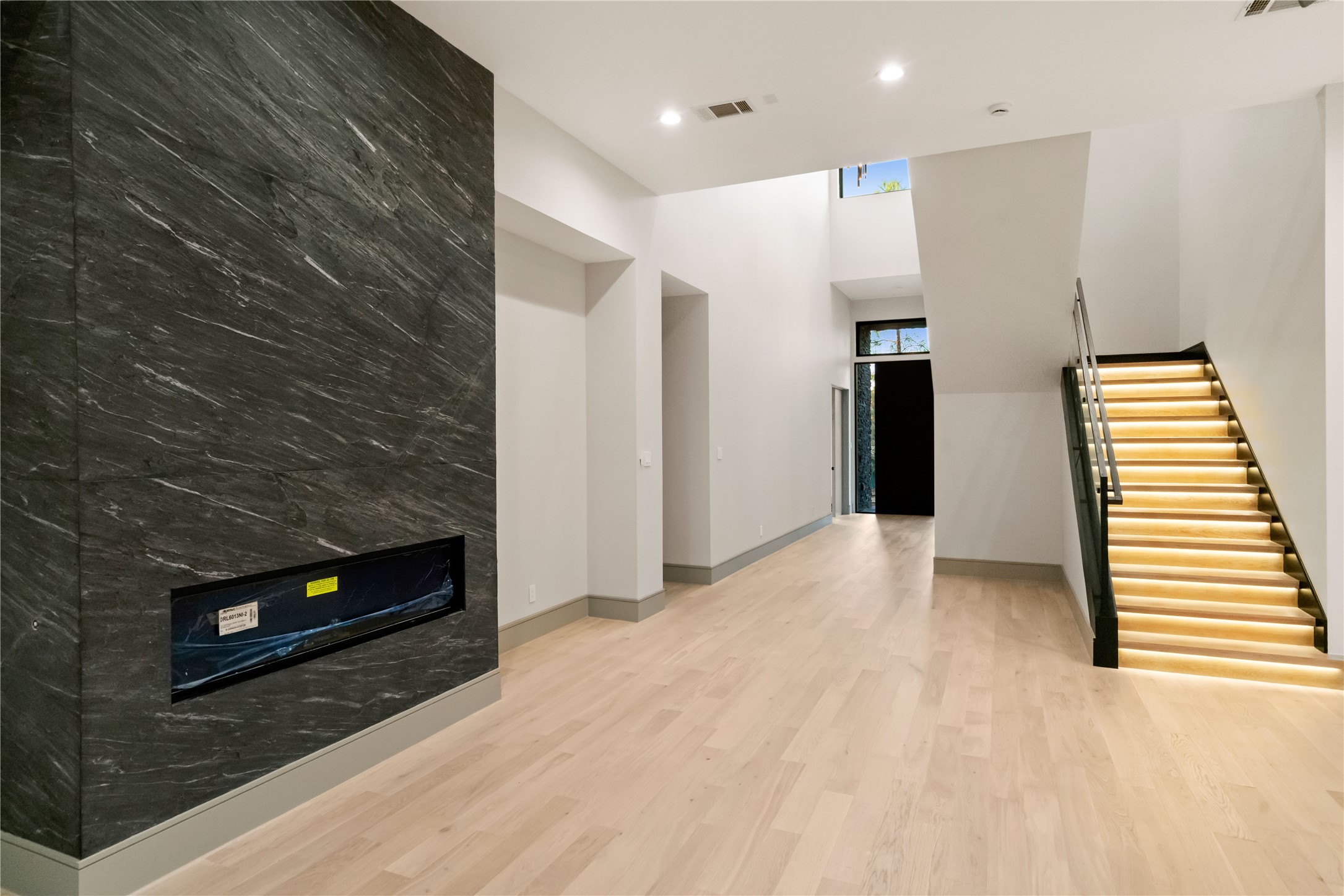 47 Pronghorn Place Spring, TX 77389 - Photo 4 of 44 This modern interior features a spacious, high-ceilinged room with light wood flooring and a sleek, dark stone accent wall with a built-in fireplace. The staircase is illuminated with contemporary lighting, enhancing the open, airy feel of the space.