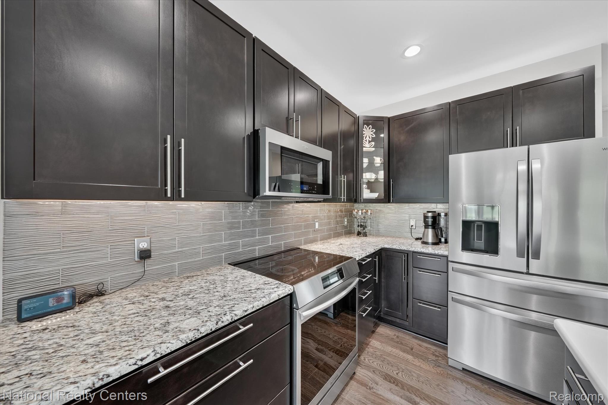 740 North Mill Street South Lyon, MI 48178 - Photo 19 of 56 Contemporary kitchen featuring dark wood cabinetry, light granite countertops, stainless steel appliances, a textured tile backsplash, and wood-finish flooring