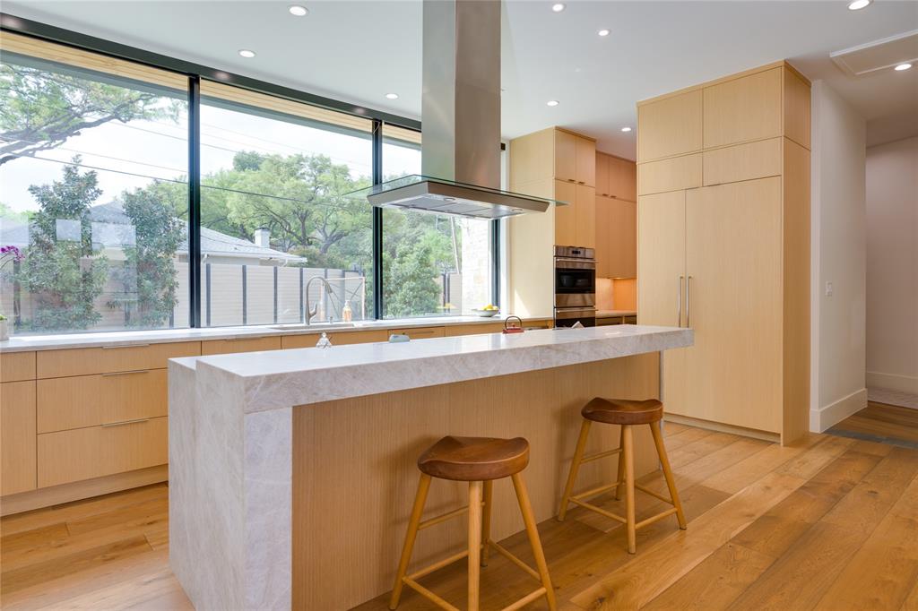 5736 Stonegate Road Dallas, TX 75209 - Photo 14 of 29 Kitchen featuring light wood-finish cabinetry, a waterfall island with a light-toned countertop, a stainless steel range hood, built-in double ovens, and expansive windows