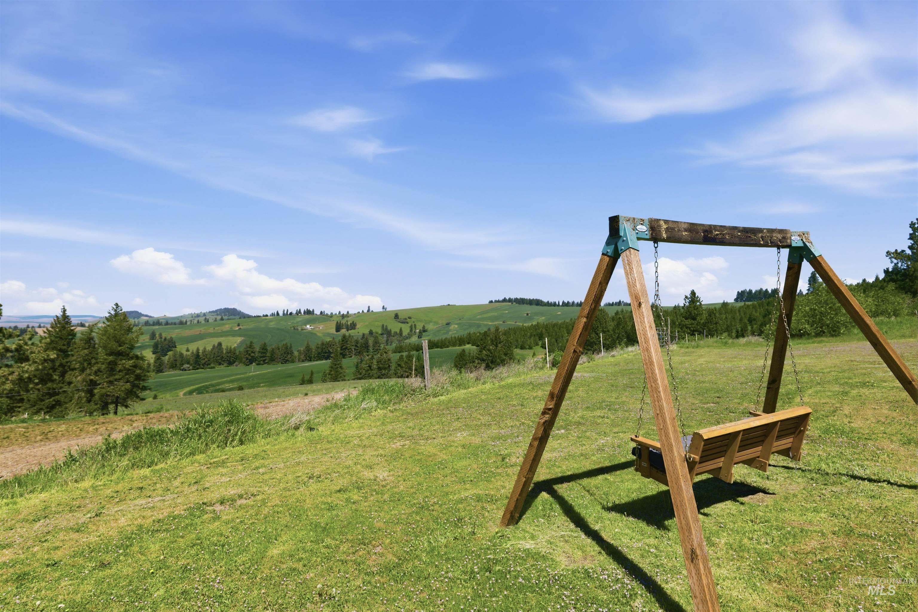 1049 Libey Road Viola, ID 83872 - Photo 23 of 43 Outdoor wooden swing with bench seating situated on a grassy area, overlooking rolling green hills and distant trees under a bright blue sky
