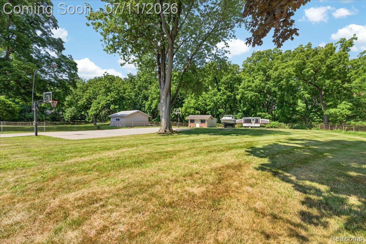 23750 East River Road Grosse Ile, MI 48138 - Photo 33 of 54 Expansive outdoor space featuring a paved sports court with basketball hoops, mature trees providing natural shade, and two utility sheds for storage