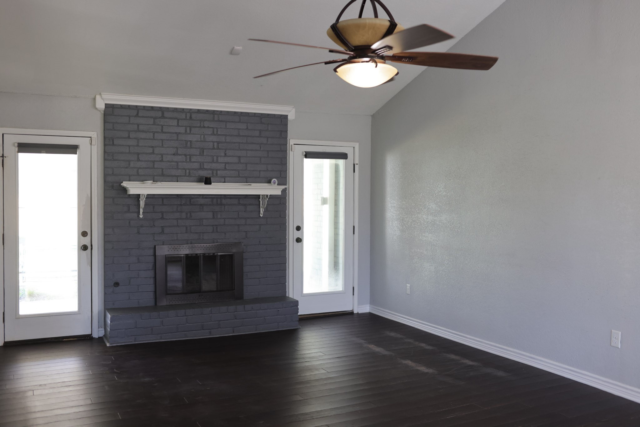 407 Tamarind Street Lake Jackson, TX 77566 - Photo 7 of 36 This room features a modern, dark brick fireplace with a mantel, flanked by two glass doors. It has dark wood flooring, light gray walls, and a ceiling fan, creating a cozy yet contemporary feel.