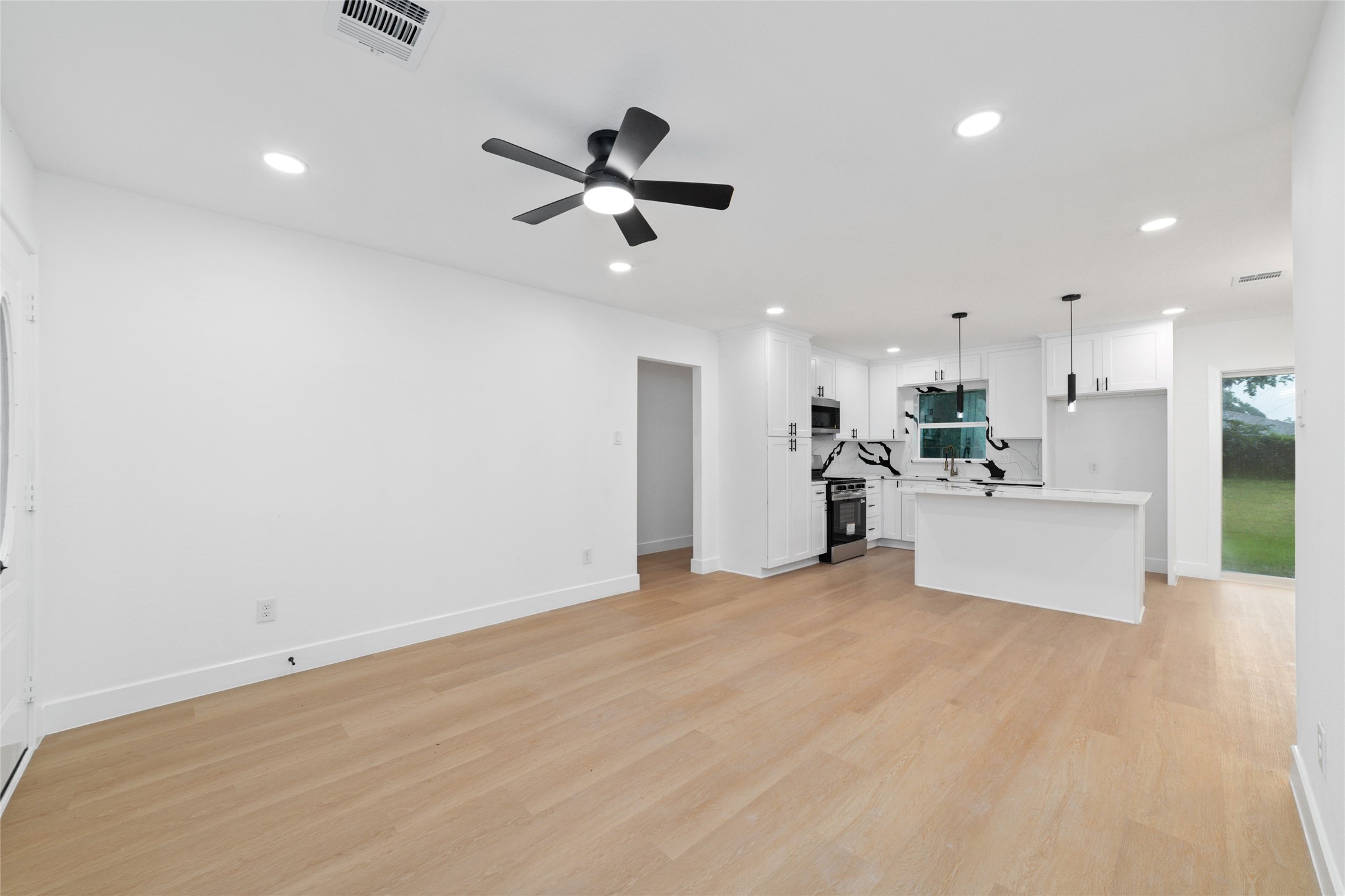 5709 Langley Road Houston, TX 77016 - Photo 5 of 27 This photo showcases a modern, open-concept living area with light wood flooring and a sleek kitchen. The kitchen features white cabinets, a marble backsplash, and an island with pendant lighting. The space is well-lit with recessed lighting and includes a ceiling fan.