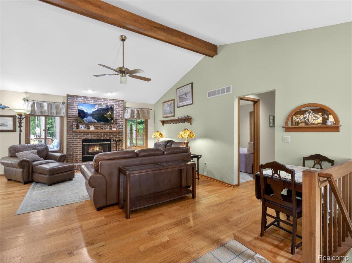 9714 Naiad Road Clarkston, MI 48348 - Photo 18 of 61 The room features a vaulted ceiling with exposed wood beams, a brick fireplace with mantel, recessed lighting, warm wood flooring, and a ceiling fan with integrated lighting for year-round comfort.