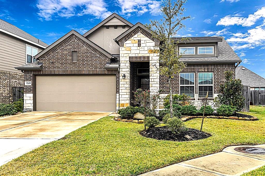 17439 Chester Valley Trail Hockley, TX 77447 - Photo 1 of 22 This photo showcases a modern, single-story brick home with a two-car garage and well-maintained landscaping. The house features a combination of brick and stone accents, a neat driveway, and a small front garden. The sky is clear, adding to the home's curb appeal.