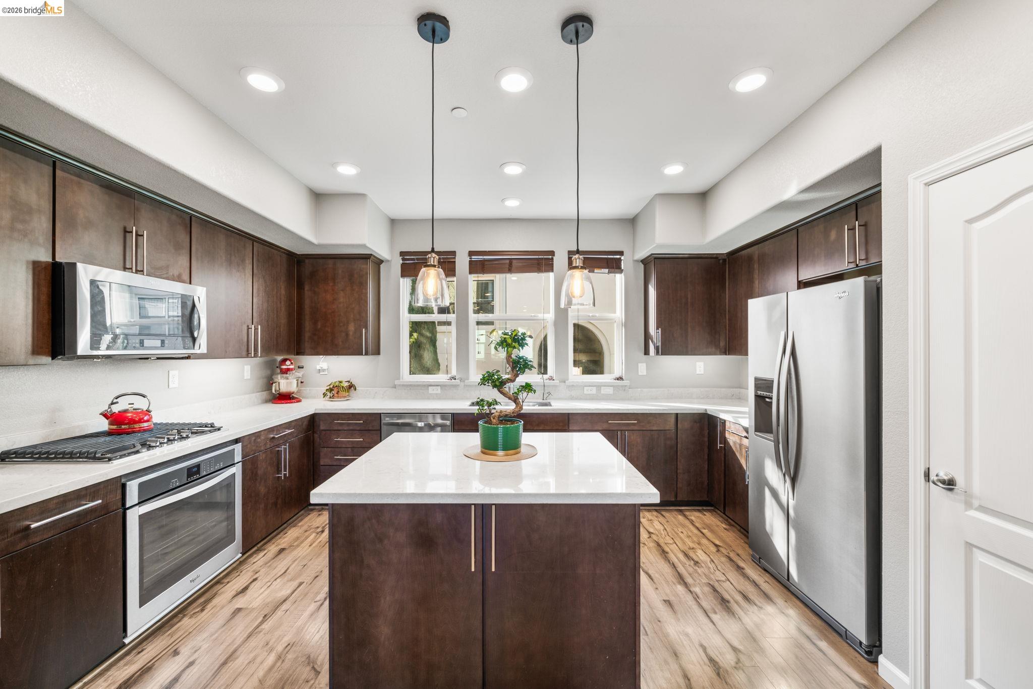 Kitchen featuring dark wood finish cabinets, stainless steel appliances, pendant lighting, a center island, and white Caesarstone quartz countertops. and laminated flooring