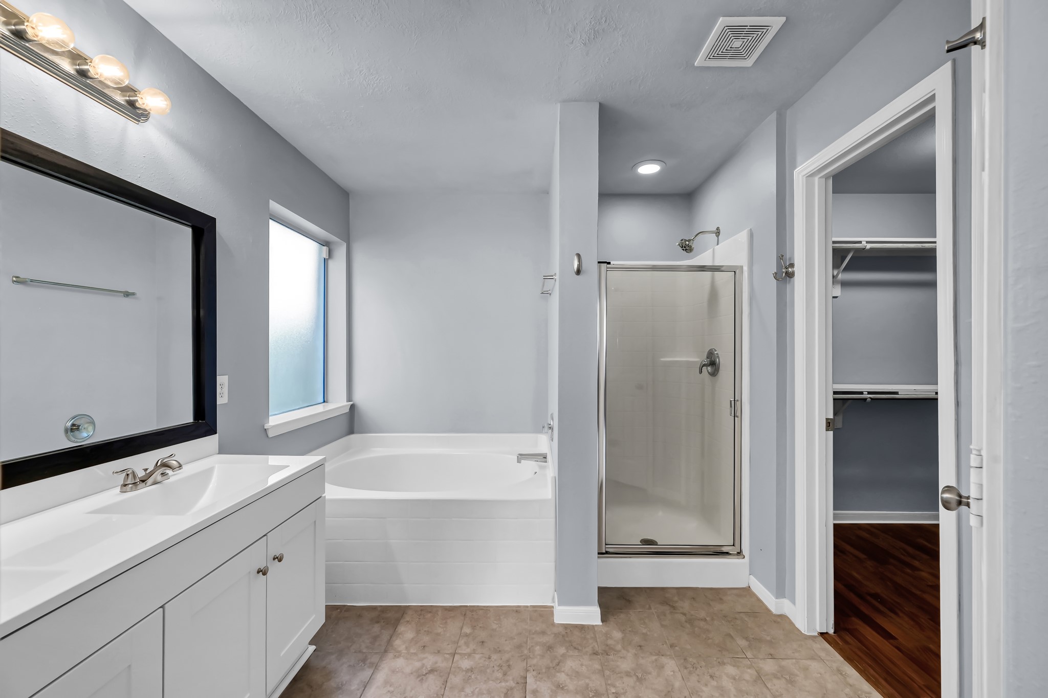 7811 Holly Berry Court Cypress, TX 77433 - Photo 13 of 24 This bathroom features a sleek design with a new modern vanity and a large mirror. It includes a spacious bathtub and a separate glass-enclosed shower. The room has ample natural light and connects to a walk-in closet, enhancing convenience and storage.