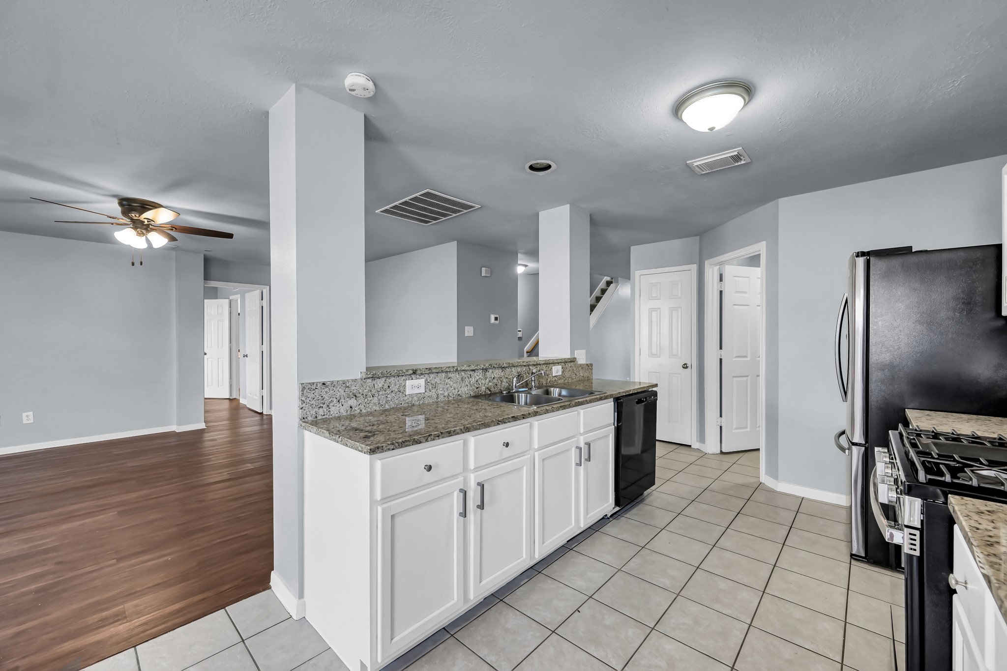 7811 Holly Berry Court Cypress, TX 77433 - Photo 9 of 24 This photo showcases a modern kitchen with granite countertops, white cabinetry, and stainless steel appliances. The open layout connects to a living area with wood flooring and a ceiling fan, providing a spacious and inviting feel.