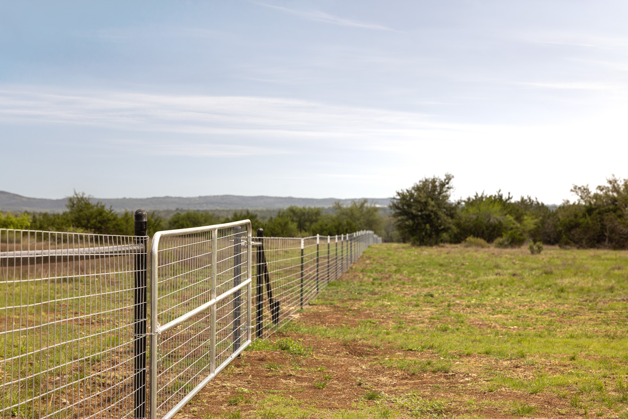 2609 Prochnow Road Dripping Springs, TX 78620 - Photo 8 of 14 Extensive perimeter fencing with a metal gate, set against a backdrop of distant hills and mature trees