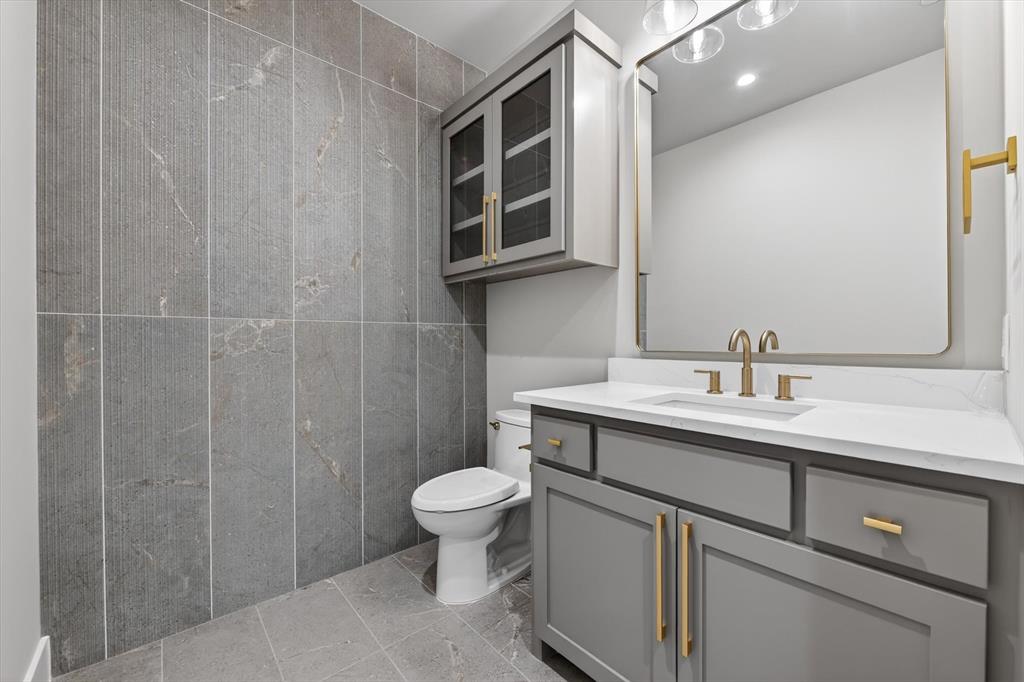 11028 Edgemere Road Dallas, TX 75230 - Photo 30 of 40 Powder room featuring full-height tiled walls, tiled flooring, a gray vanity with gold-tone hardware, a white countertop, and a rectangular mirror with a gold-tone frame