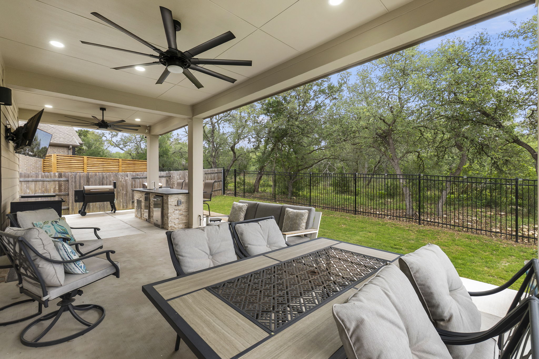 460 Constitution Way Kyle, TX 78640 - Photo 34 of 40 Covered patio featuring a built-in outdoor kitchen with stone veneer and a counter surface, two ceiling fans, recessed lighting, and a metal fence bordering a natural treeline