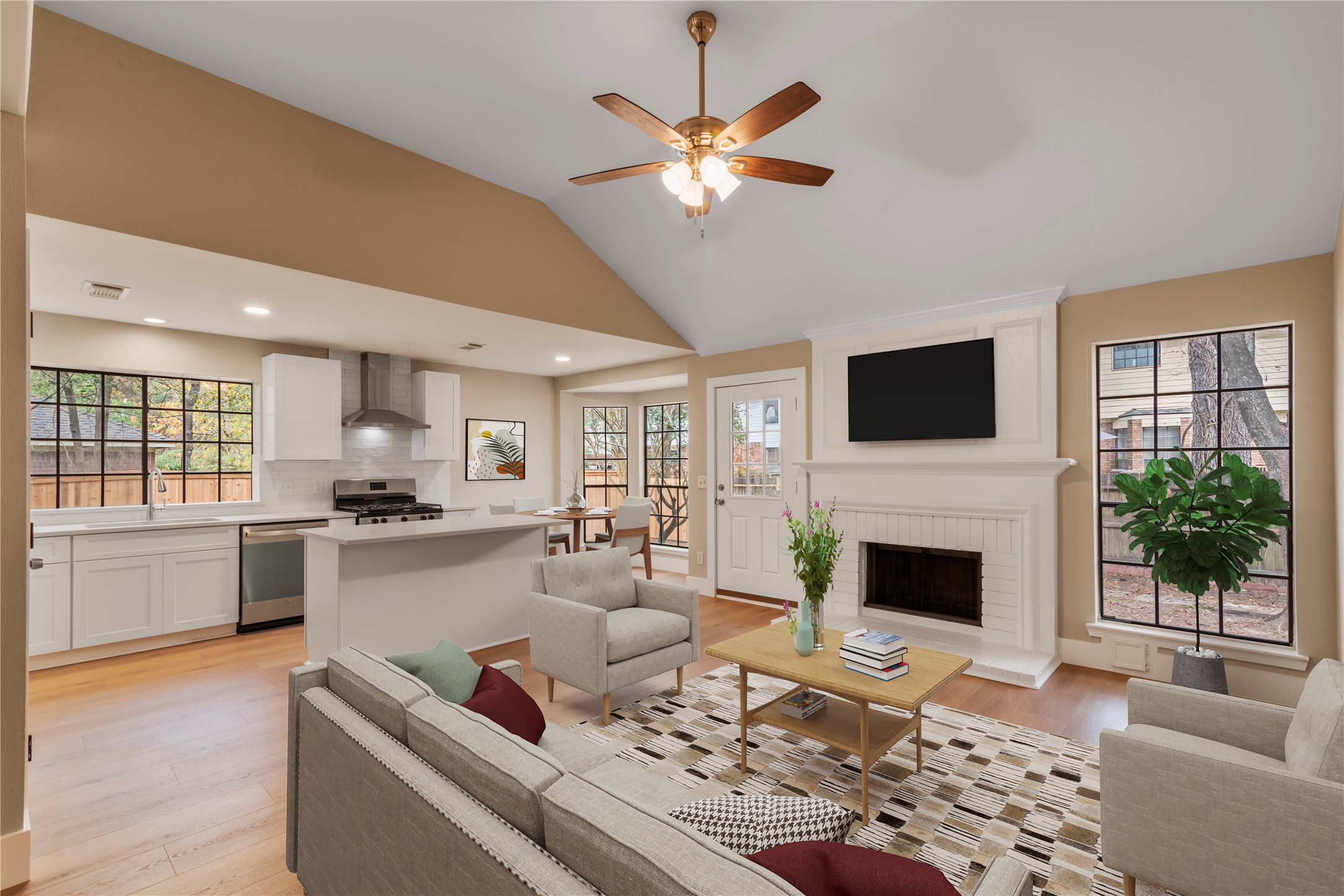10 White Fawn Drive Spring, TX 77381 - Photo 6 of 25 This photo showcases a bright, open-concept living area featuring a modern kitchen with white cabinetry, a central island, and stainless steel appliances. The adjoining living room has a cozy fireplace, ceiling fan, and large windows, offering a warm and inviting space for relaxation. Virtually staged.