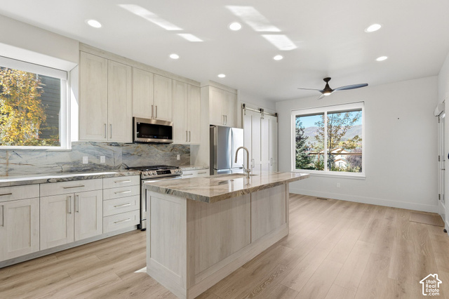 Modern kitchen featuring light wood-finish cabinetry, stainless steel appliances, a large island with a sink, stone countertops, and wood-finish flooring