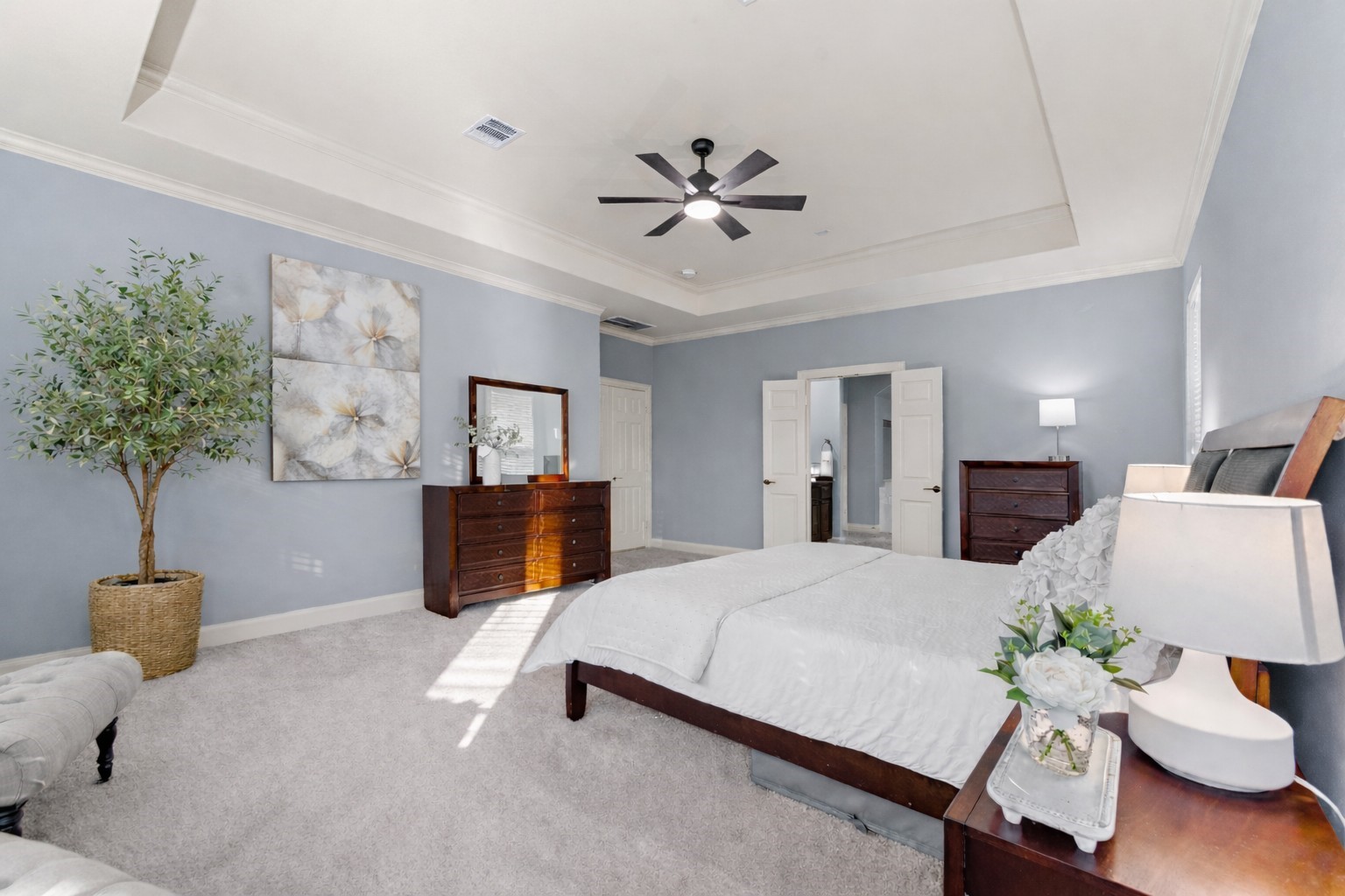 70 Valera Ridge Drive Spring, TX 77389 - Photo 23 of 37 This spacious bedroom features soft blue walls, a tray ceiling with a modern fan, and elegant decor. It includes a comfortable bed, two wooden dressers, and ample natural light, creating a serene and inviting atmosphere.