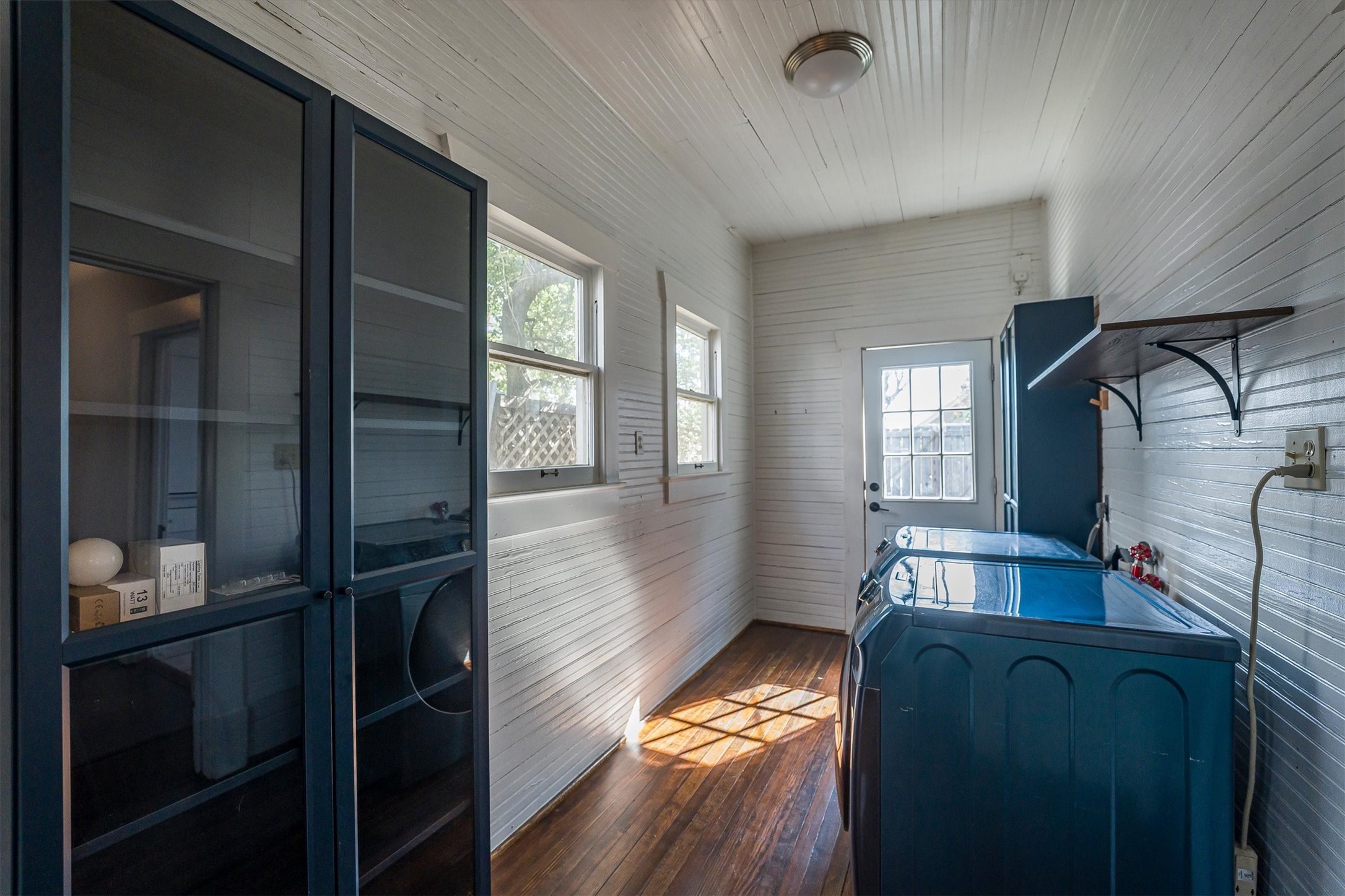 210 Tabor Street Houston, TX 77009 - Photo 11 of 18 This photo shows a bright laundry room with white paneled walls and a wooden floor. It features a washer and dryer, built-in shelving, and storage cabinets. A window and a glass door allow natural light to fill the space. The backdoor leads to the backyard.