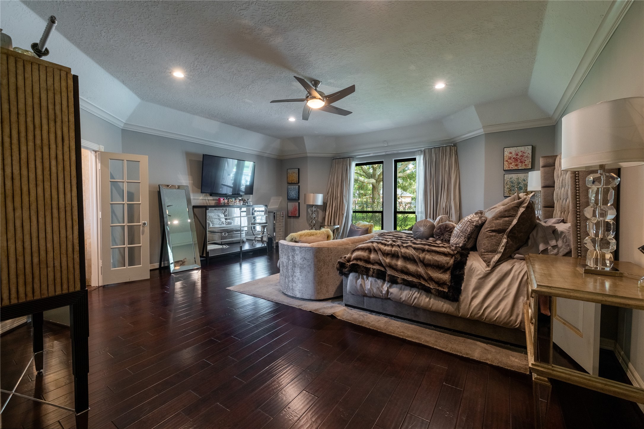 4002 Silver Ridge Boulevard Missouri City, TX 77459 - Photo 18 of 50 This is a cozy, Primary bedroom featuring rich, dark hardwood floors and a vaulted ceiling with a ceiling fan. The room has ample natural light from the large windows with a view of greenery. It's furnished with a large bed, elegant bedding, a mirrored dresser, and a mounted TV for comfort and entertainment.