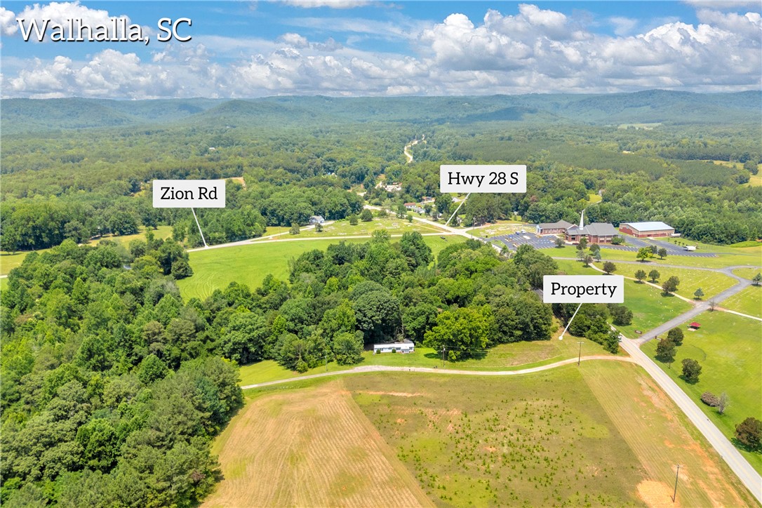 An aerial perspective reveals the property's advantageous placement between two roads amidst lush greenery and distant hills.