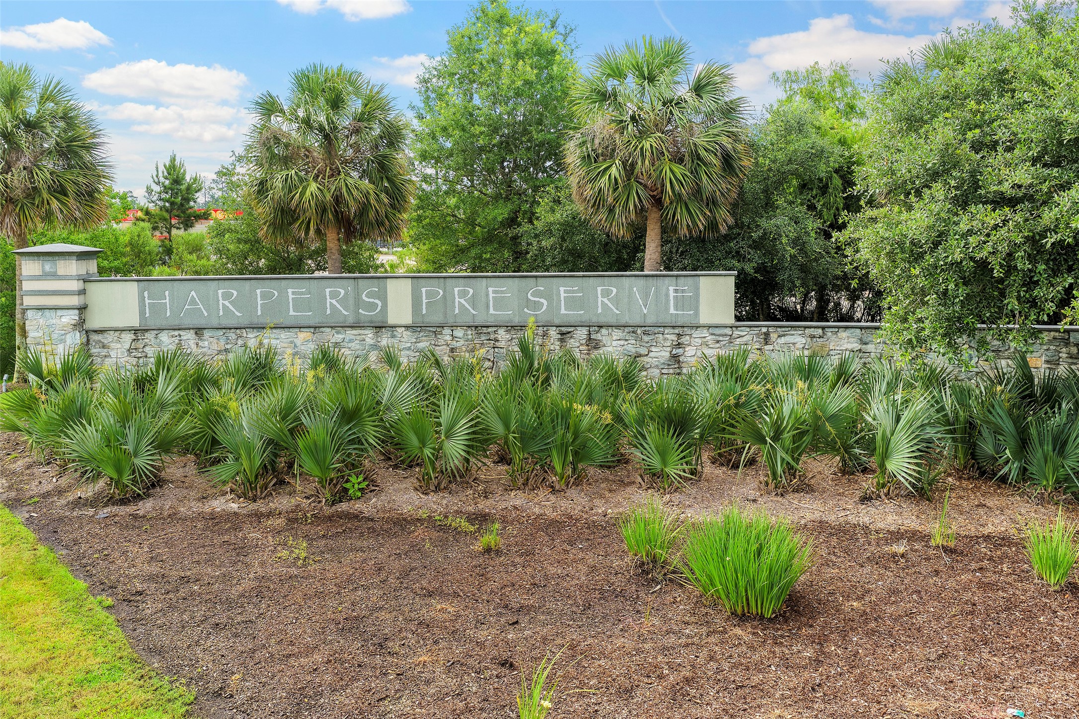 10603 Whitejacket Conroe, TX 77385 - Photo 32 of 42 Entrance to Harper's Preserve, featuring a stone wall with the community name and lush landscaping, including palm trees and greenery, set under a bright blue sky.