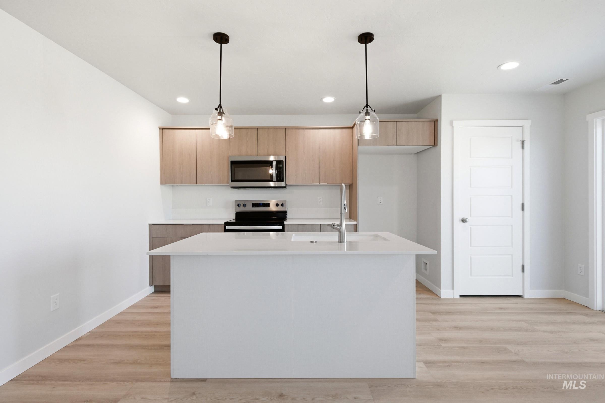 10794 Rutland Street Caldwell, ID 83605 - Photo 2 of 22 Contemporary kitchen featuring a large central island with an integrated sink, light wood-finish cabinetry, stainless steel appliances, and sleek pendant lighting