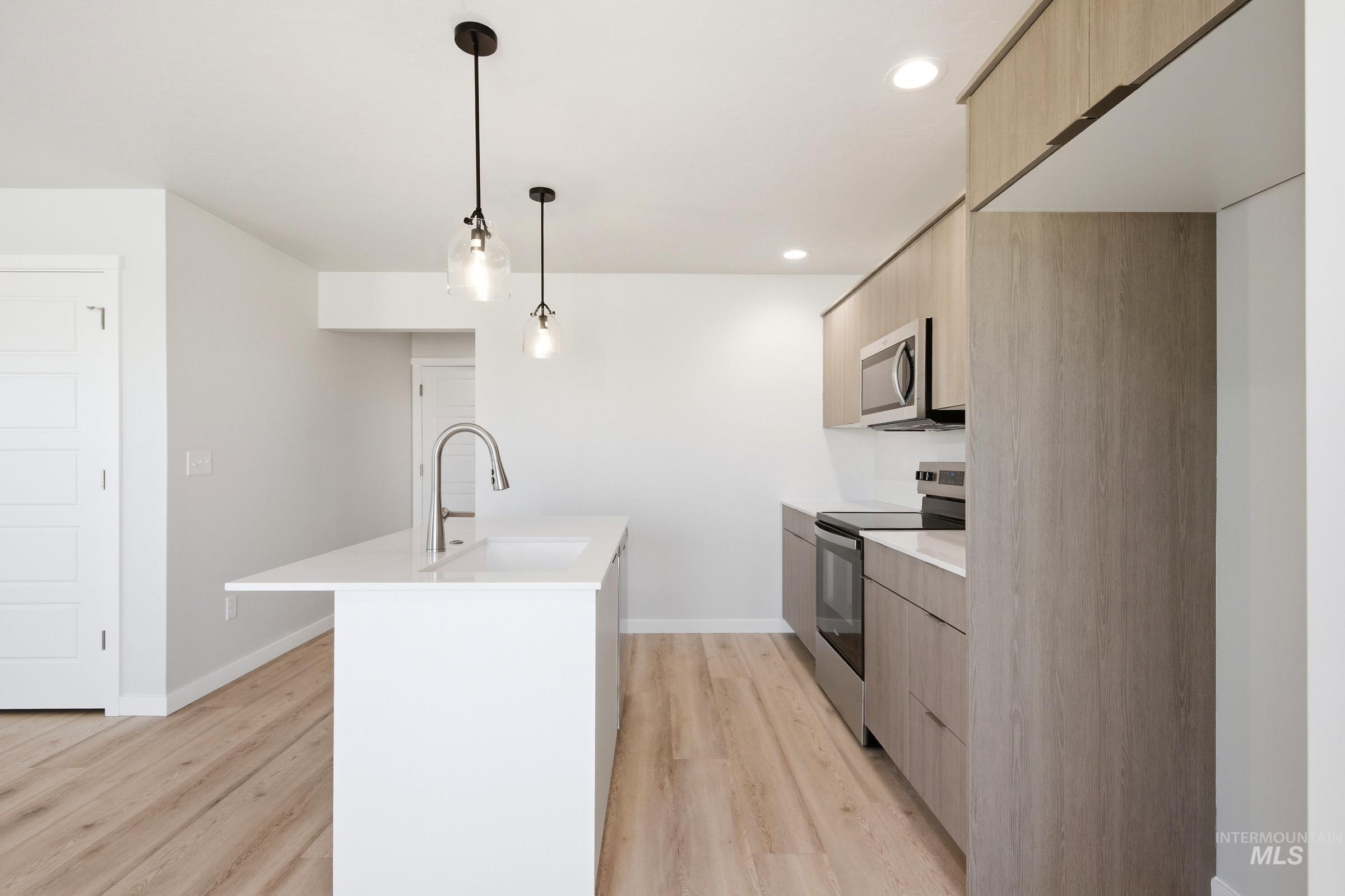 10794 Rutland Street Caldwell, ID 83605 - Photo 7 of 22 Open-concept kitchen featuring a white island with an undermount sink, pendant lighting, stainless steel appliances, wood-finish flooring, and light wood-grain cabinetry