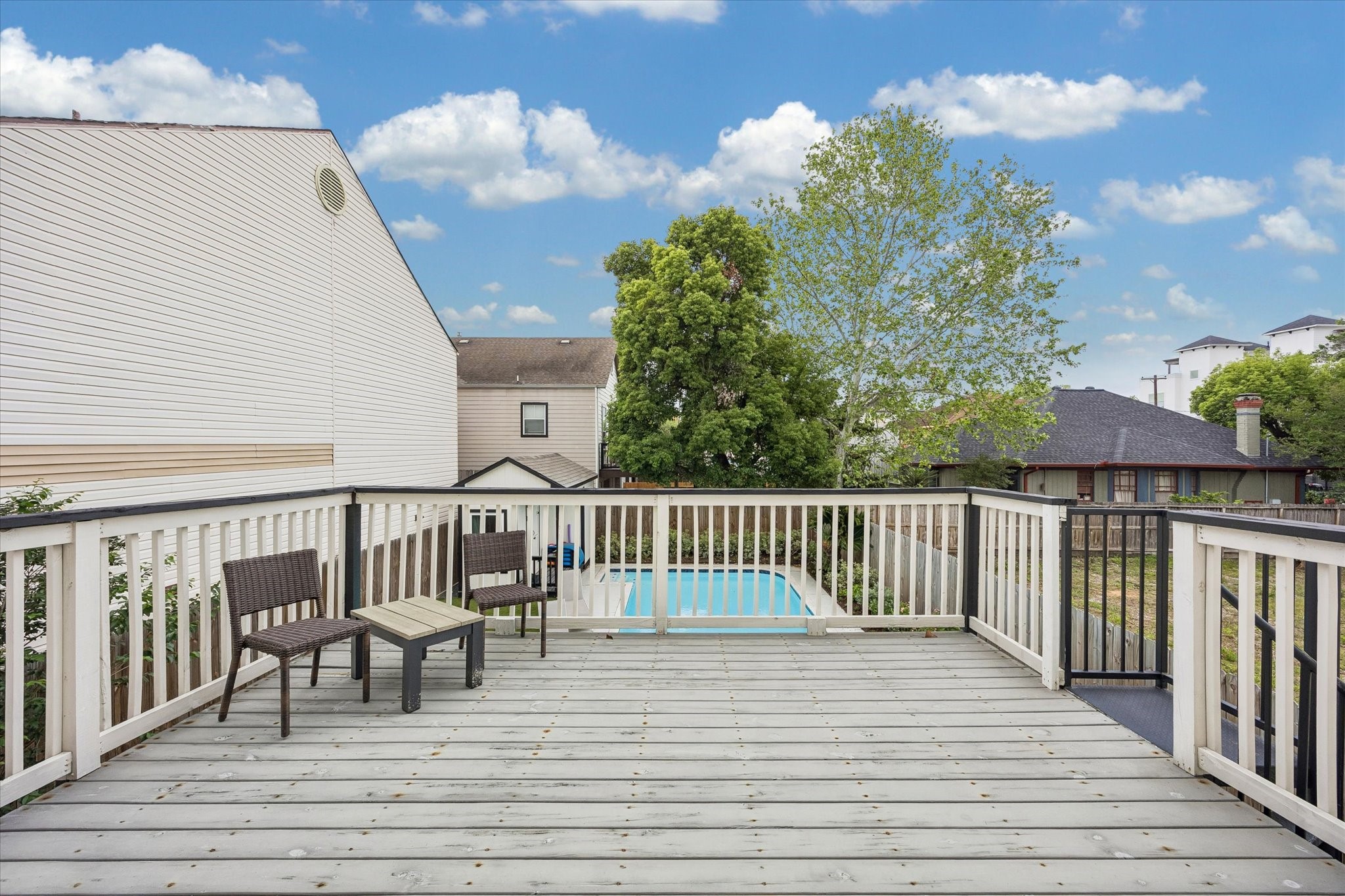 1420 Indiana Street Houston, TX 77006 - Photo 2 of 31 This photo features a spacious wooden deck, overlooking a backyard with a pool. The area is enclosed by a fence, with neighboring houses and lush trees in the background, under a partly cloudy sky.