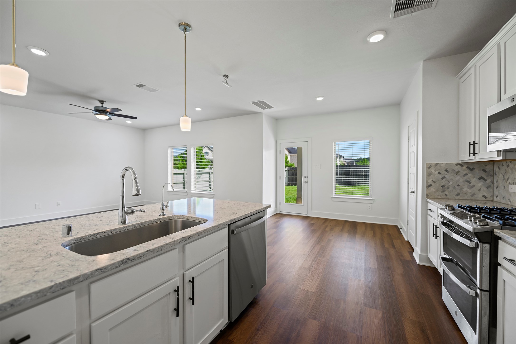 10603 Whitejacket Conroe, TX 77385 - Photo 26 of 42 This modern kitchen features a sleek island with a built-in sink, stainless steel appliances, and white cabinetry. It opens to a bright living area with wood-style flooring and access to a backyard through a glass door.