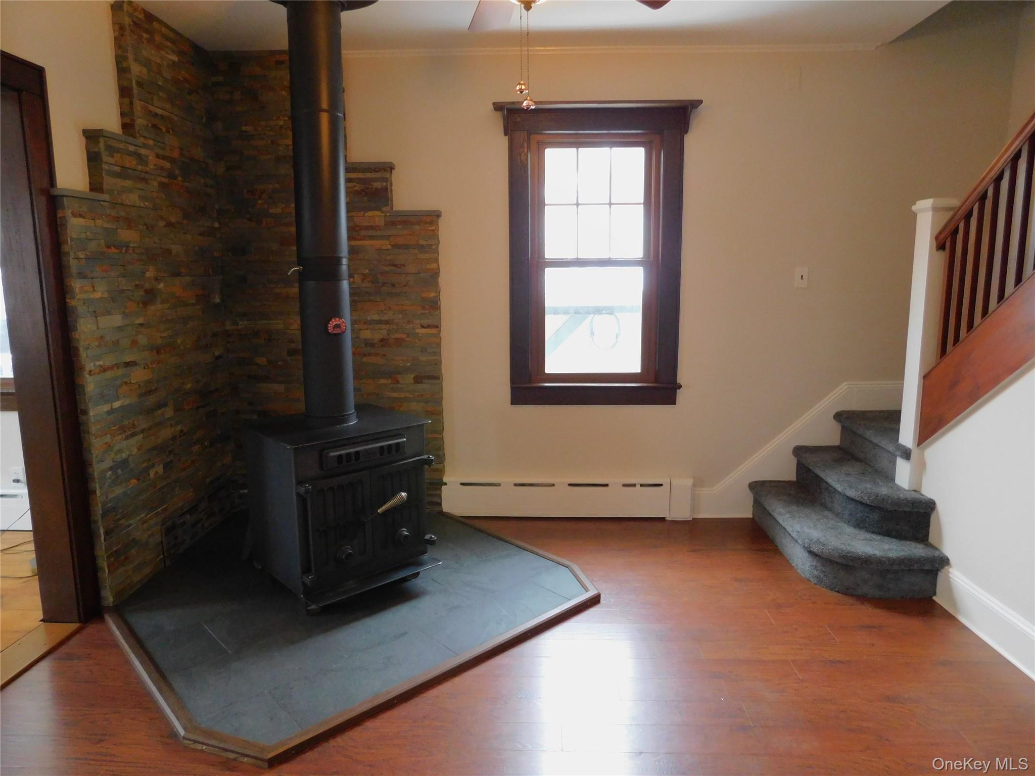 84 Cedar Road Wading River, NY 11792 - Photo 9 of 17 Den/ living area with a wood stove, wood finished floors, a baseboard radiator, stairway, and crown molding