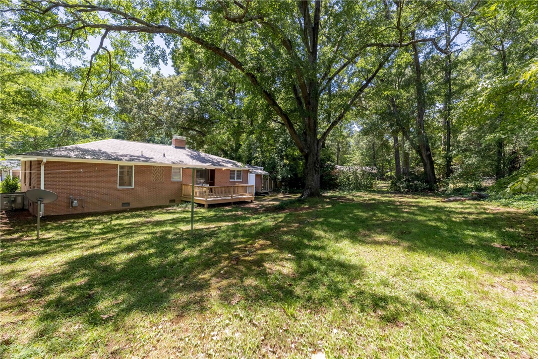 207 Brook Street Clemson, SC 29631 - Photo 28 of 39 A spacious backyard with mature trees surrounds a charming brick home featuring a wooden deck, perfect for outdoor relaxation.
