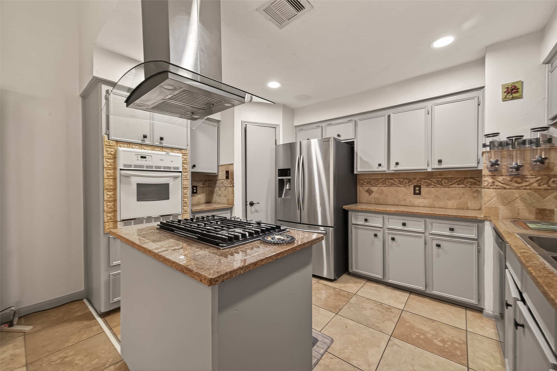 11331 Oakcenter Drive Houston, TX 77072 - Photo 8 of 35 This kitchen features modern stainless steel appliances, including a built-in oven and refrigerator. It has tiled floors, ample cabinet space, and a central island with a gas cooktop and overhead range hood. The countertops are complemented by decorative backsplash tiles, creating a stylish and functional cooking space.