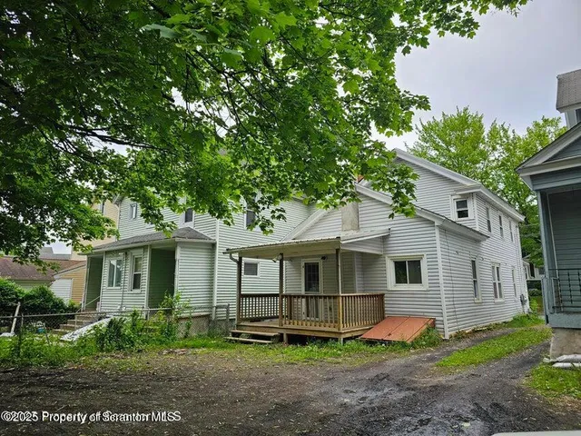 a view of a white house next to a yard with potted plants and large trees