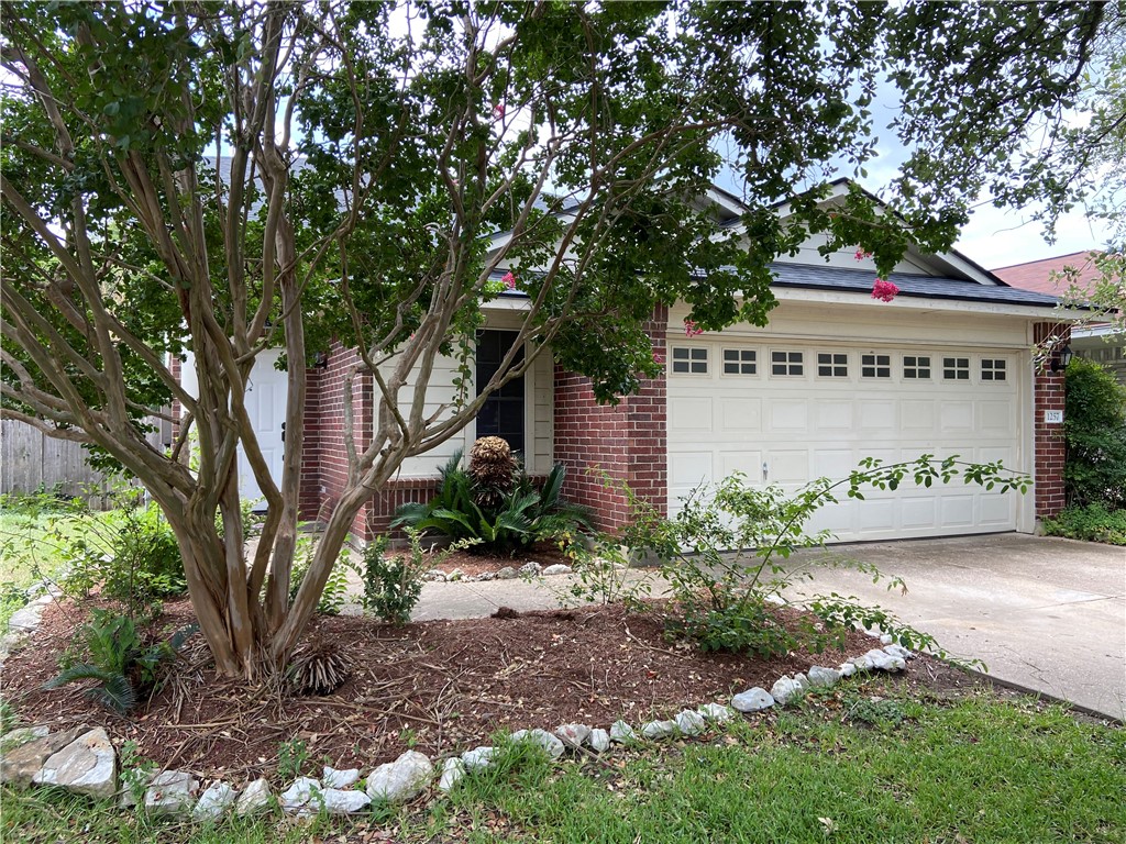 Street View of property hidden behind natural elements with brick siding, an attached garage, and driveway