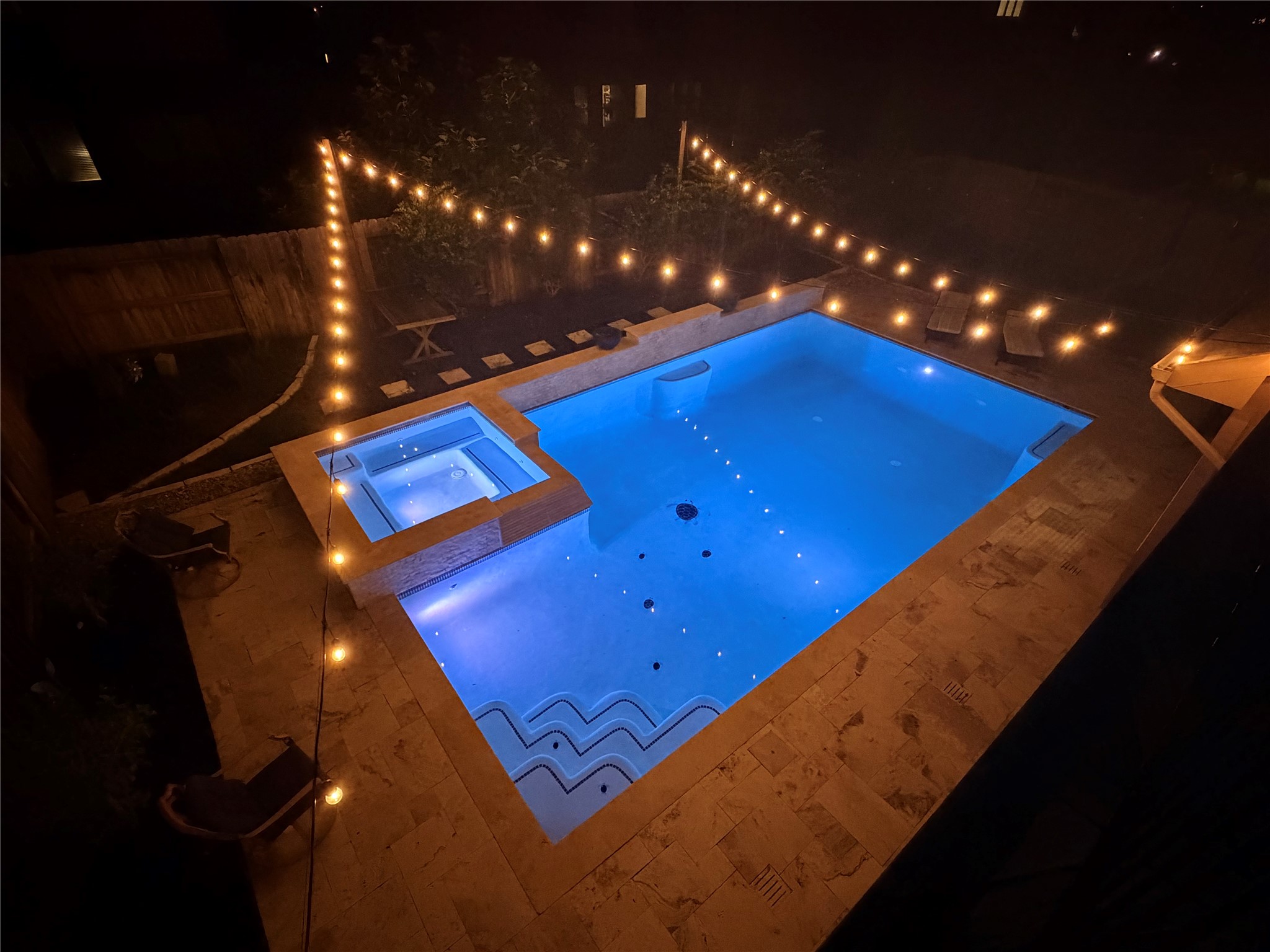 25611 Ellis Ridge Court Katy, TX 77494 - Photo 42 of 49 This inviting backyard features a modern swimming pool and a hot tub, surrounded by elegant stone tiles. Soft string lights create a warm, ambient glow, perfect for evening relaxation or entertaining.