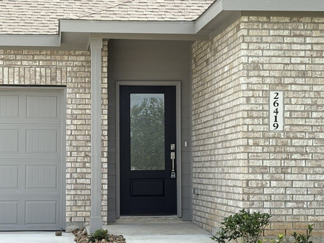 26419 Virginia Wild Rye Lane Katy, TX 77493 - Photo 5 of 31 This photo shows a modern brick home exterior with a covered entryway. It features a dark front door with glass paneling, adjacent to a garage with a matching gray door. The house number is clearly displayed on the wall, and there are small shrubs along the walkway.