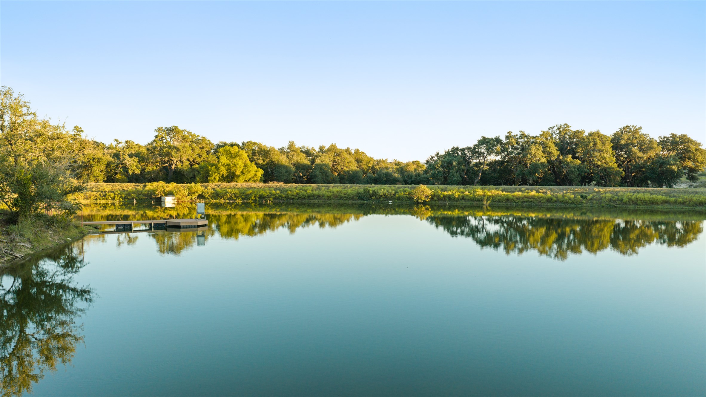 4318 Maenius Road Blanco, TX 78606 - Photo 1 of 11 A tranquil private pond mirrors the surrounding mature trees under a clear sky.