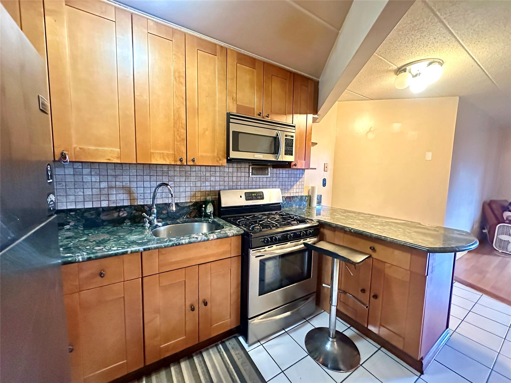 130-14 60th Avenue, Unit 2E Queens, NY 11355 - Photo 5 of 8 Kitchen featuring stainless steel appliances, a peninsula, decorative backsplash, light tile patterned flooring, and dark stone countertops