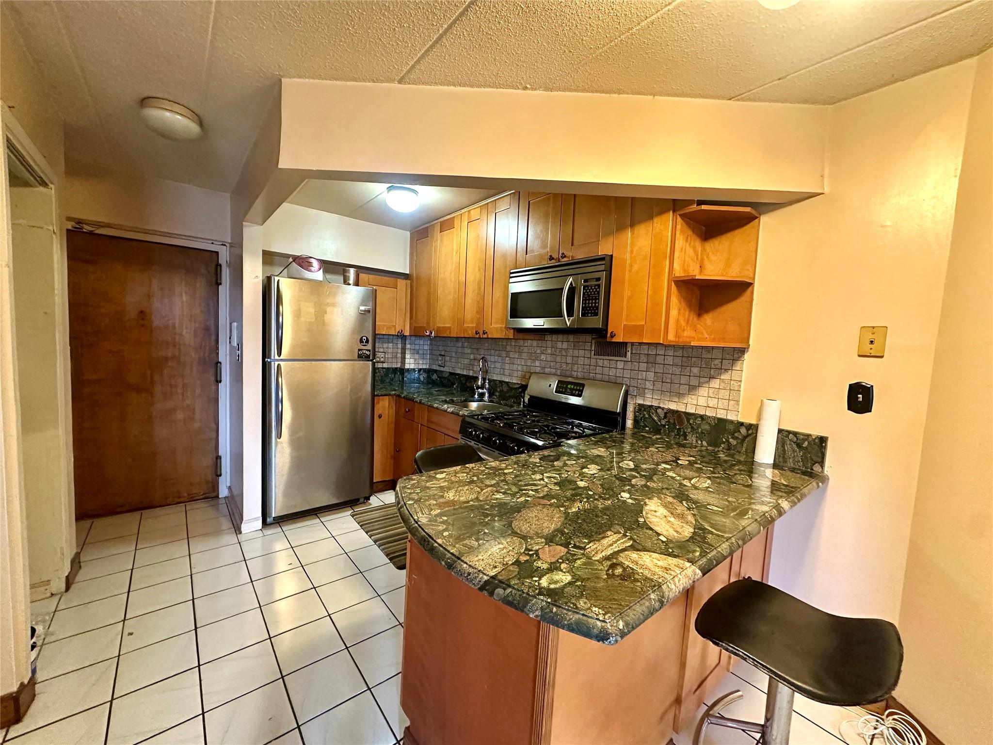 130-14 60th Avenue, Unit 2E Queens, NY 11355 - Photo 8 of 8 Kitchen with stainless steel appliances, a peninsula, decorative backsplash, brown cabinets, and light tile patterned flooring