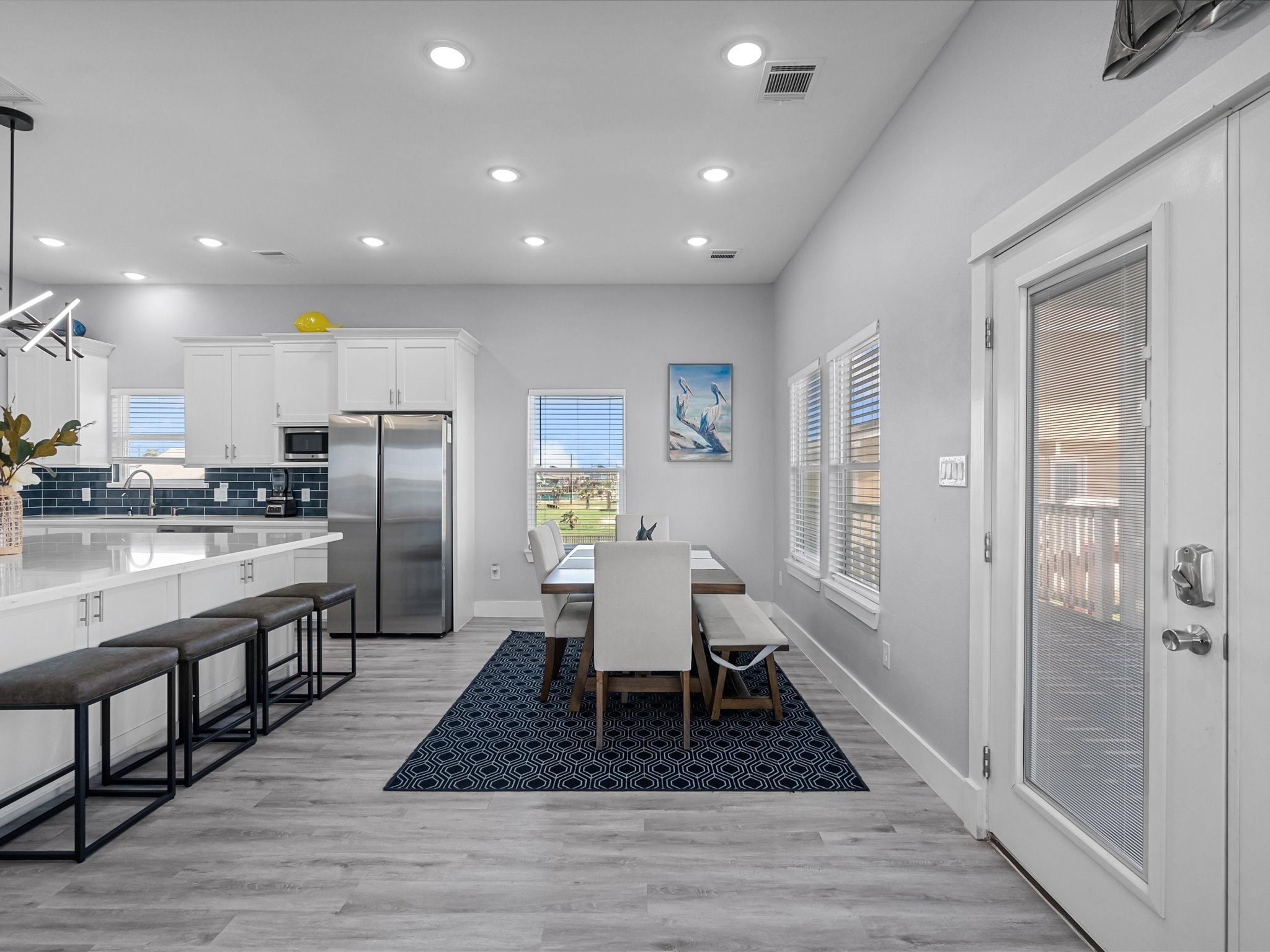 718 Beach Drive Surfside Beach, TX 77541 - Photo 12 of 49 Discover the perfect blend of style and function in this modern kitchen and dining area, featuring sleek white cabinetry, a large island with seating, stainless steel appliances, and a striking blue tile backsplash. The open layout, enhanced by stylish lighting and wood flooring, creates a bright, inviting atmosphere.