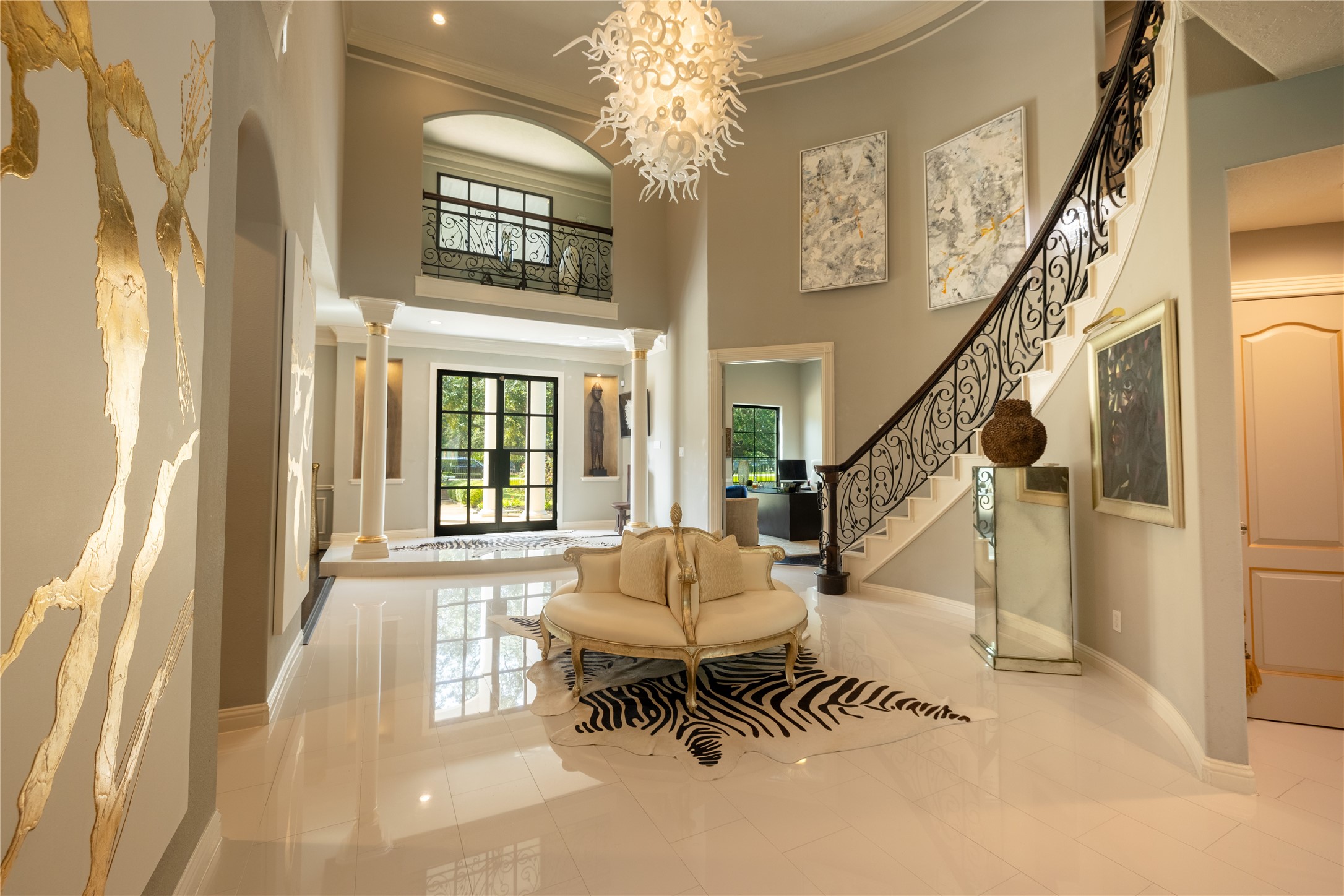 4002 Silver Ridge Boulevard Missouri City, TX 77459 - Photo 6 of 50 This is a spacious, elegant entryway featuring high ceilings with a modern chandelier, polished tile floors, and a grand staircase with ornate wrought-iron railings. The area is well-lit with natural light from large windows and is adorned with artistic decor and columns, presenting a luxurious and welcoming ambiance.
