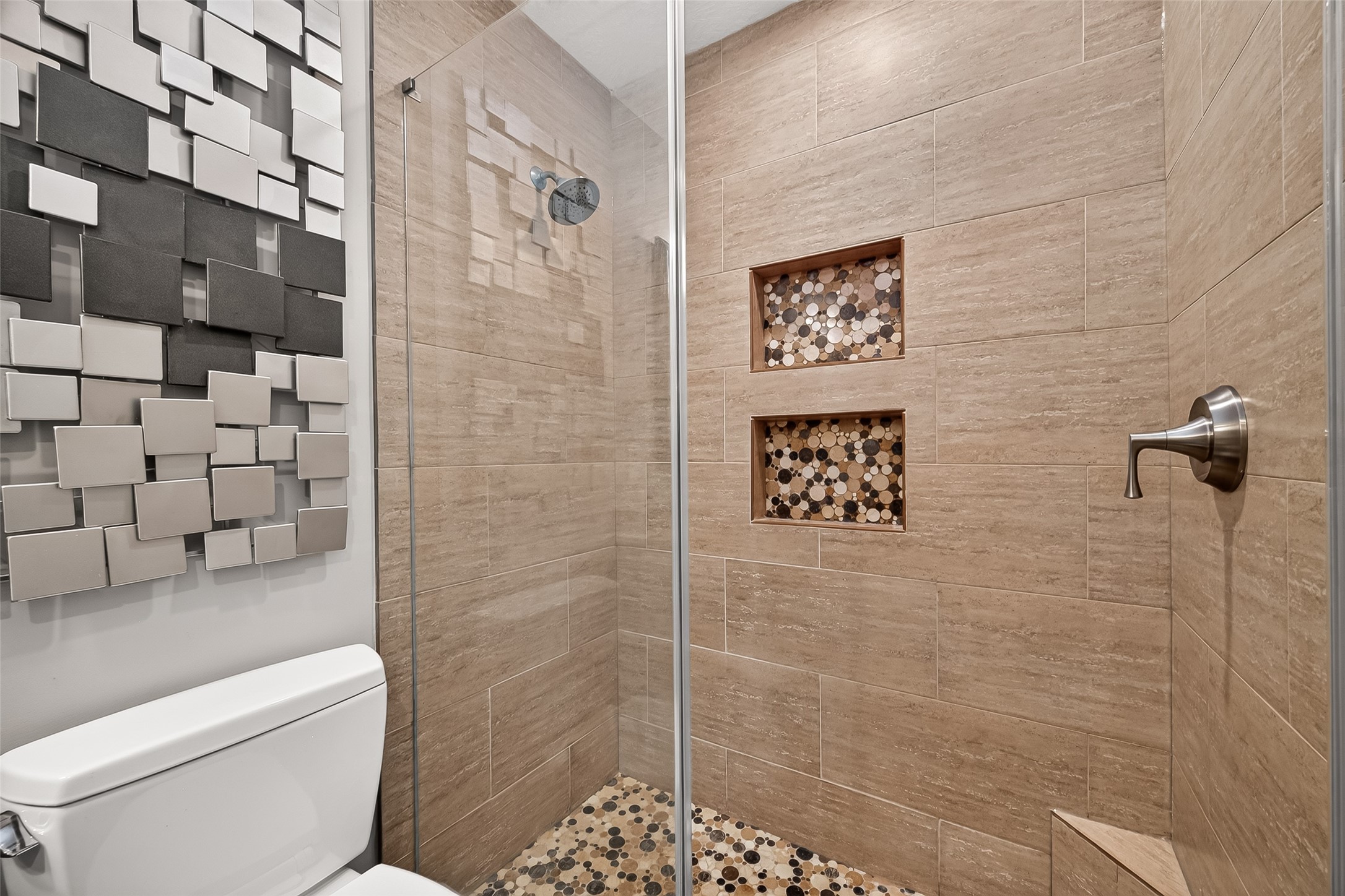 24719 Wilderness Road Spring, TX 77380 - Photo 33 of 42 Modern primary bathroom with a stylish walk-in shower featuring beige tiles and mosaic accents. It includes a sleek showerhead and built-in niches. A decorative wall panel adds a contemporary touch.