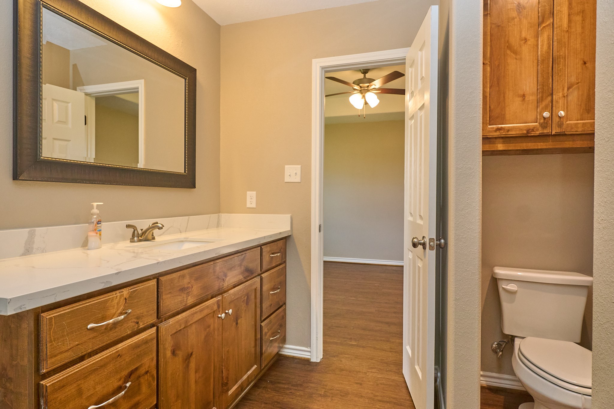 1807 Nuches Lane Bryan, TX 77803 - Photo 17 of 27 This photo features a modern bathroom with a wooden vanity, marble countertop, and large mirror. It includes a toilet and has a doorway leading to a room with a ceiling fan and wooden flooring. The neutral color palette gives a warm, inviting feel.