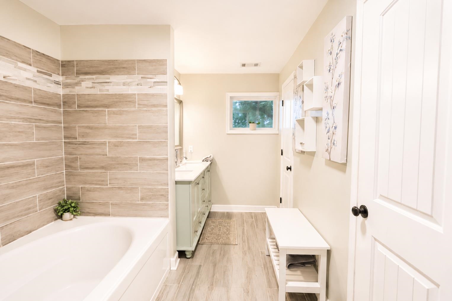 10093 Mt Carmel Road Brighton, TN 38011 - Photo 30 of 36 Bathroom featuring a white soaking tub with wood-look tile surround and mosaic accent band, a double vanity with light blue cabinetry and dual sinks, wood-finish flooring, a window, and white paneled doors