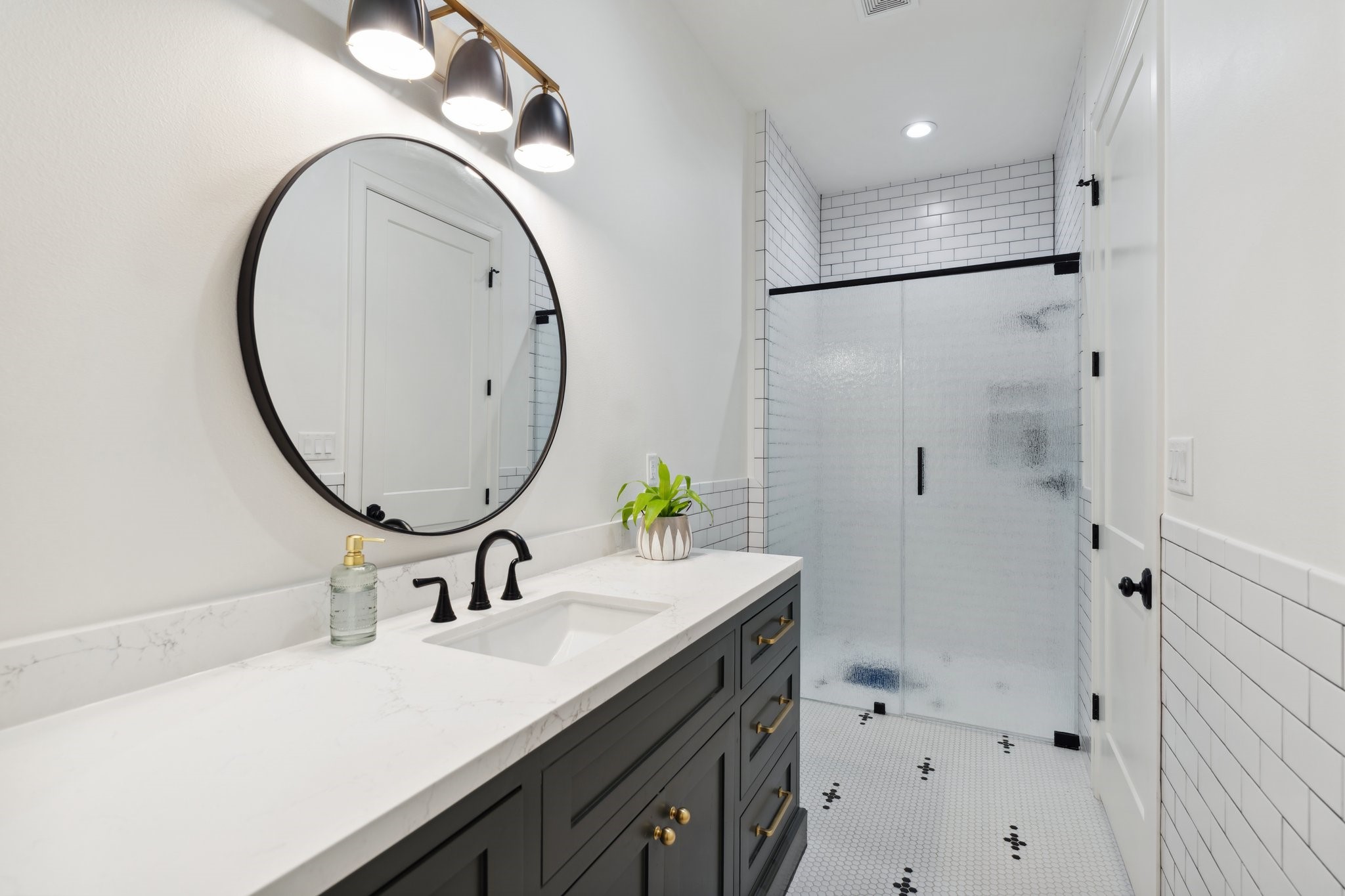4052 Kuykendall Road Bellville, TX 77418 - Photo 23 of 40 This bathroom features a modern design with a sleek black vanity, white marble countertop, and a round mirror. It has a spacious shower with frosted glass doors and stylish black fixtures, complemented by white subway tile walls and hexagonal floor tiles.
