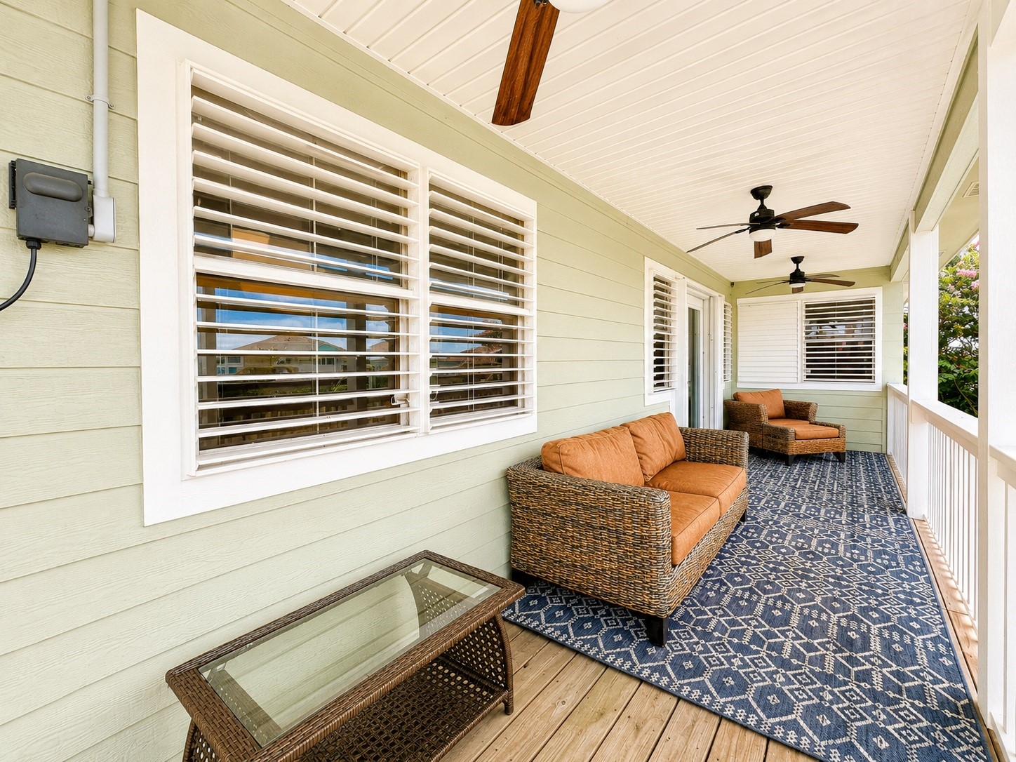 135 Sand Shoals Road Freeport, TX 77541 - Photo 18 of 20 This photo showcases a spacious and inviting covered porch with light green siding, featuring comfortable wicker furniture with orange cushions, a patterned blue rug, and ceiling fans for added comfort. Perfect for relaxing or entertaining.