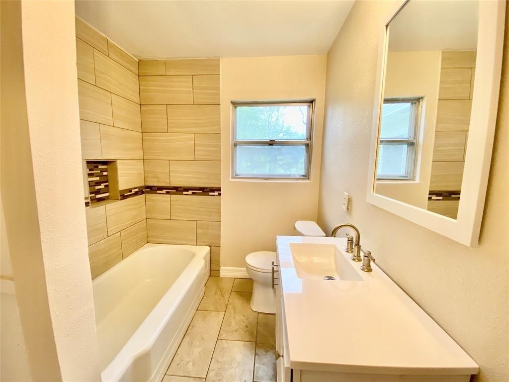 6716 Sjolander Road Baytown, TX 77521 - Photo 11 of 26 Remodeled modern bathroom featuring a white bathtub with beige tile surround, a single sink vanity, and a toilet. Natural light comes from a window above the tub. The space has a clean and bright aesthetic.