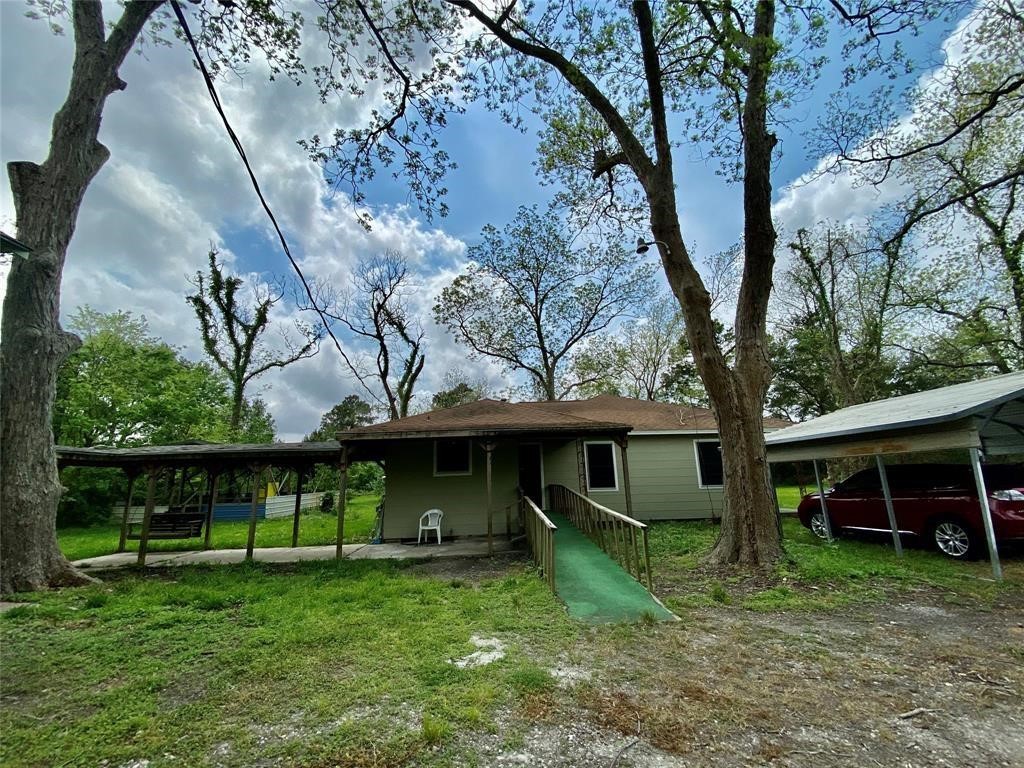 6716 Sjolander Road Baytown, TX 77521 - Photo 19 of 26 This is a single-story home with a green roof and beige siding, featuring a carport on the right and a covered porch area on the left. The property is surrounded by mature trees and has a natural, grassy yard.